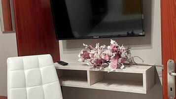 A modern bathroom with a white cushioned chair, a vanity with a mirror, a television mounted above a shelf, and a floral arrangement.