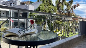 Une table ronde en verre, sur laquelle se trouvent un livre, des lunettes et une tasse, est placée sur un balcon doté d’une rambarde en métal, offrant une vue sur un quartier résidentiel avec des arbres et des immeubles.