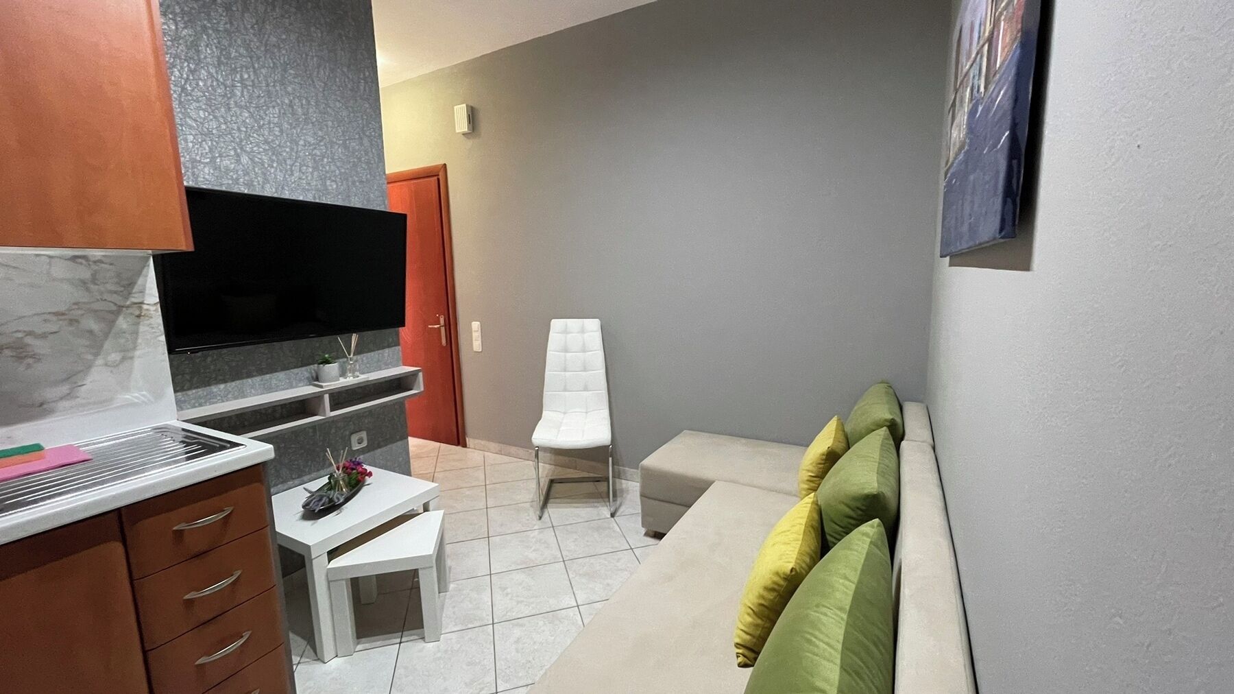 A compact living space with a TV mounted on the wall, a small white table, a white chair, and a sofa with cushions.