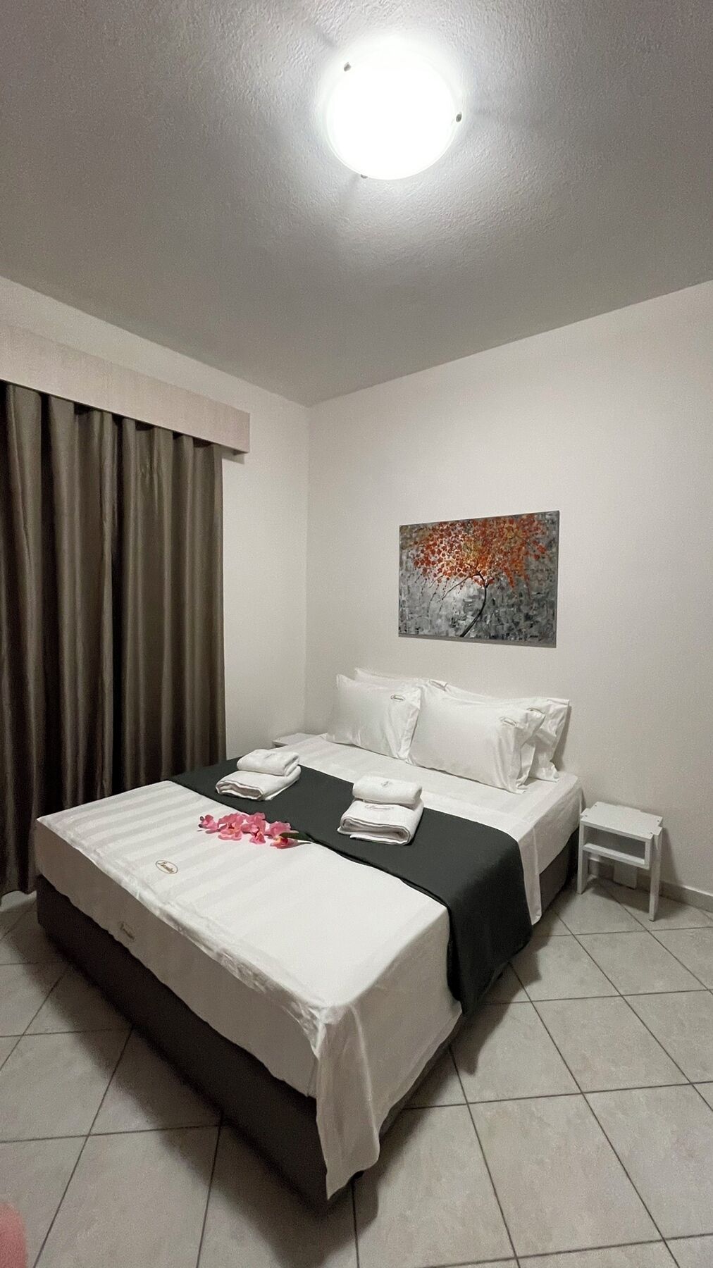 Una habitación de hotel con cama, mesita de noche, un cuadro en la pared y una ventana con cortinas.