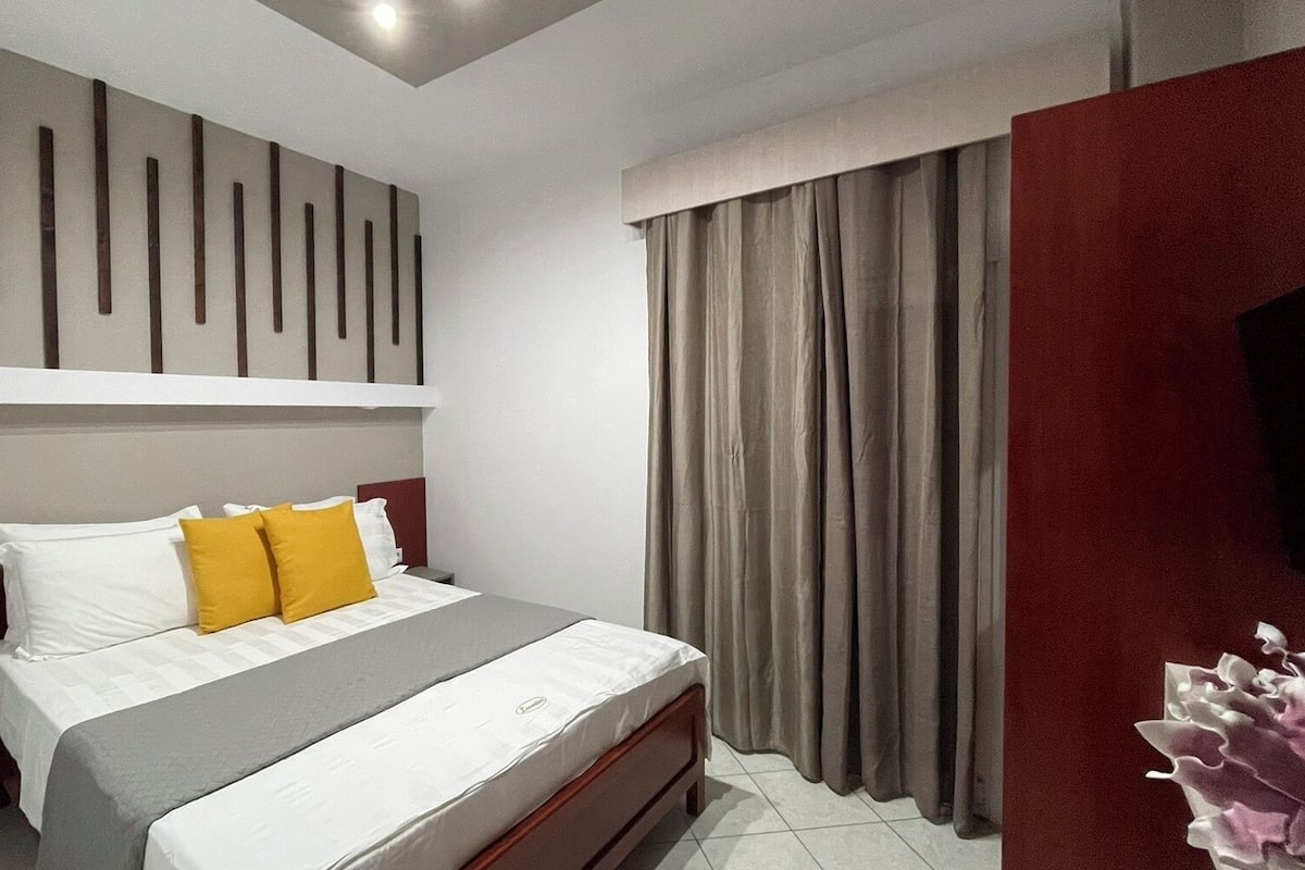 Een moderne hotelkamer met een bed, nachtkastje, ingebouwde televisie en een groot raam met gordijnen.