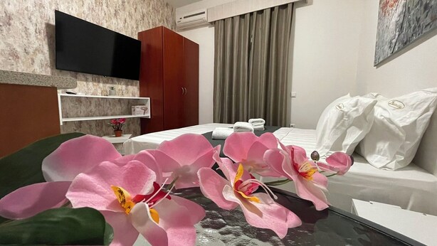 Une chambre d’hôtel moderne dotée d’un téléviseur à écran plat fixé au mur, d’un canapé avec des coussins blancs, d’une table basse en verre ornée d’orchidées roses et d’une fenêtre avec des rideaux.