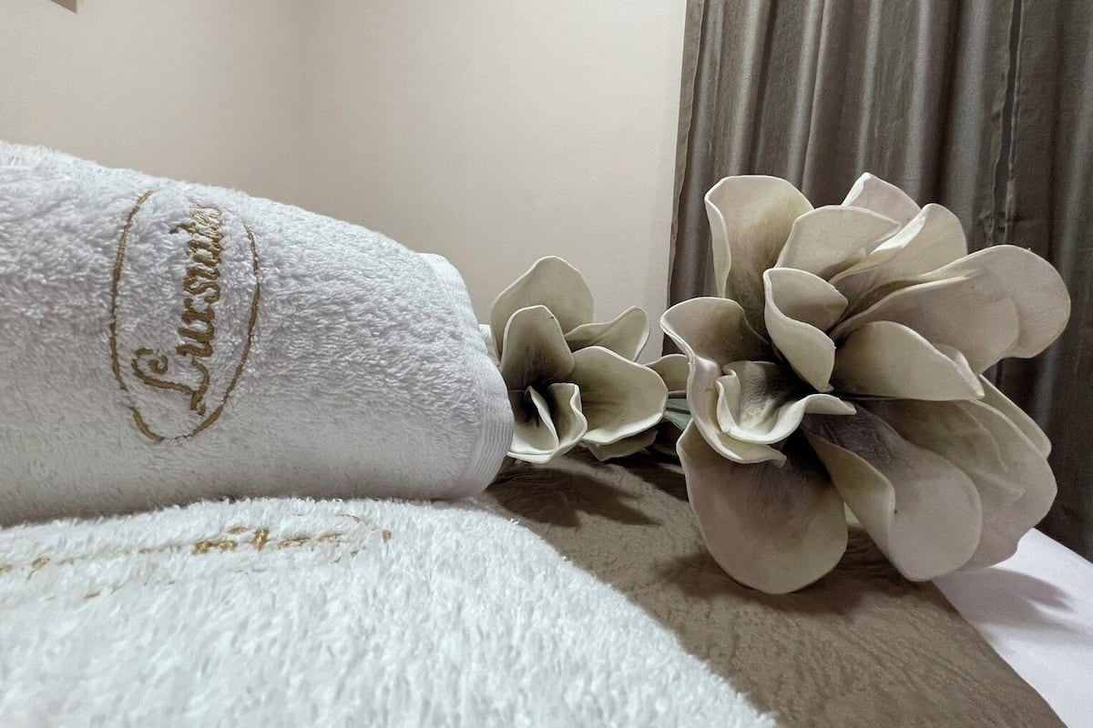 Een witte handdoek met het woord 'Lush' in goud geborduurd, geplaatst op een bed met een decoratieve bloemenstelling ernaast.