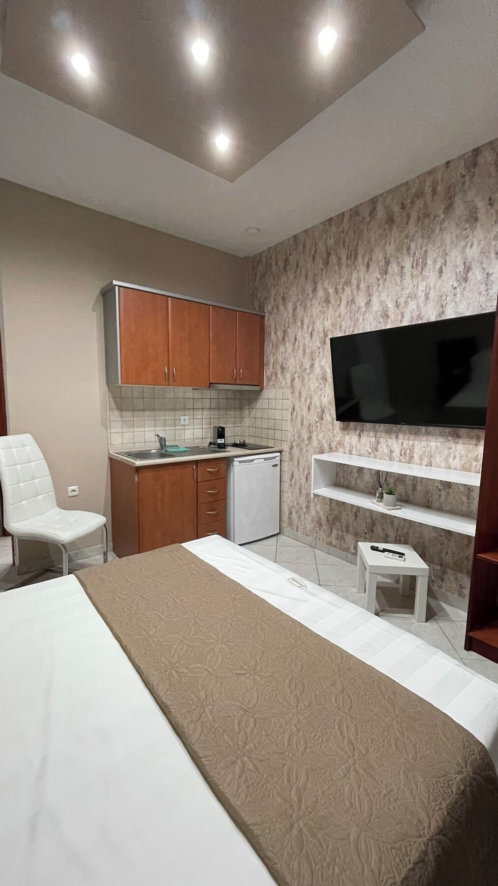 A compact bedroom with a bed, a small kitchenette, a TV mounted on the wall, and a white chair.