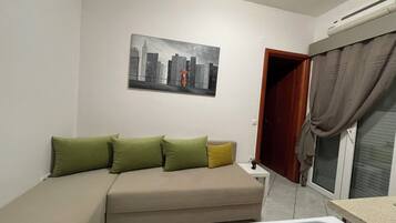 Una habitación pequeña y ordenada con un sofá, un cuadro de un paisaje urbano y una ventana con cortinas.
