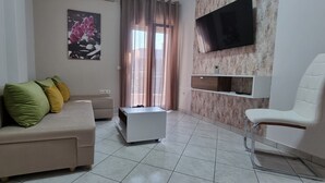 Un salon avec un canapé beige, un fauteuil blanc, une petite table d’appoint blanche, un téléviseur fixé au mur et une fenêtre avec des rideaux.