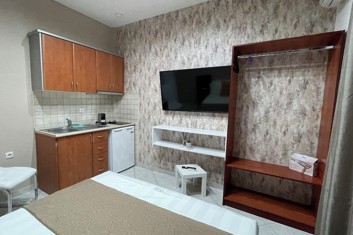 Een kleine keuken met een vaatwasser, een witte stoel, een bed, een aan de muur bevestigde televisie en een douchegedeelte.