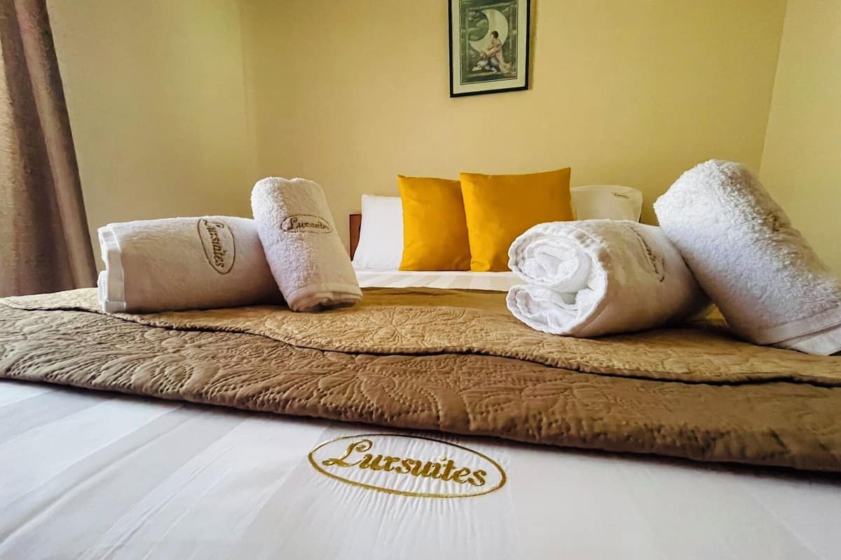 Een net opgemaakt bed met een bruin sprei, witte lakens en twee opgerolde handdoeken met de merknaam 'Luxurantes'. Ook zichtbaar is een geel kussen en een ingelijst foto.