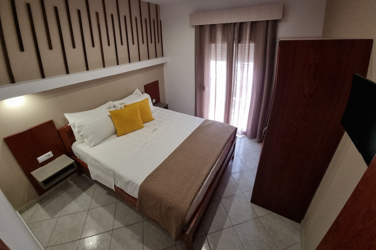Een hotelkamer met een bed, nachtkastje, kledingkast en raam met gordijnen.