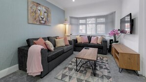 A living room with two dark grey sofas, a wooden coffee table, a pink blanket, a painting of a lion, a pink lamp, a TV, and a window with blinds. - Grindleford House (Birmingham)