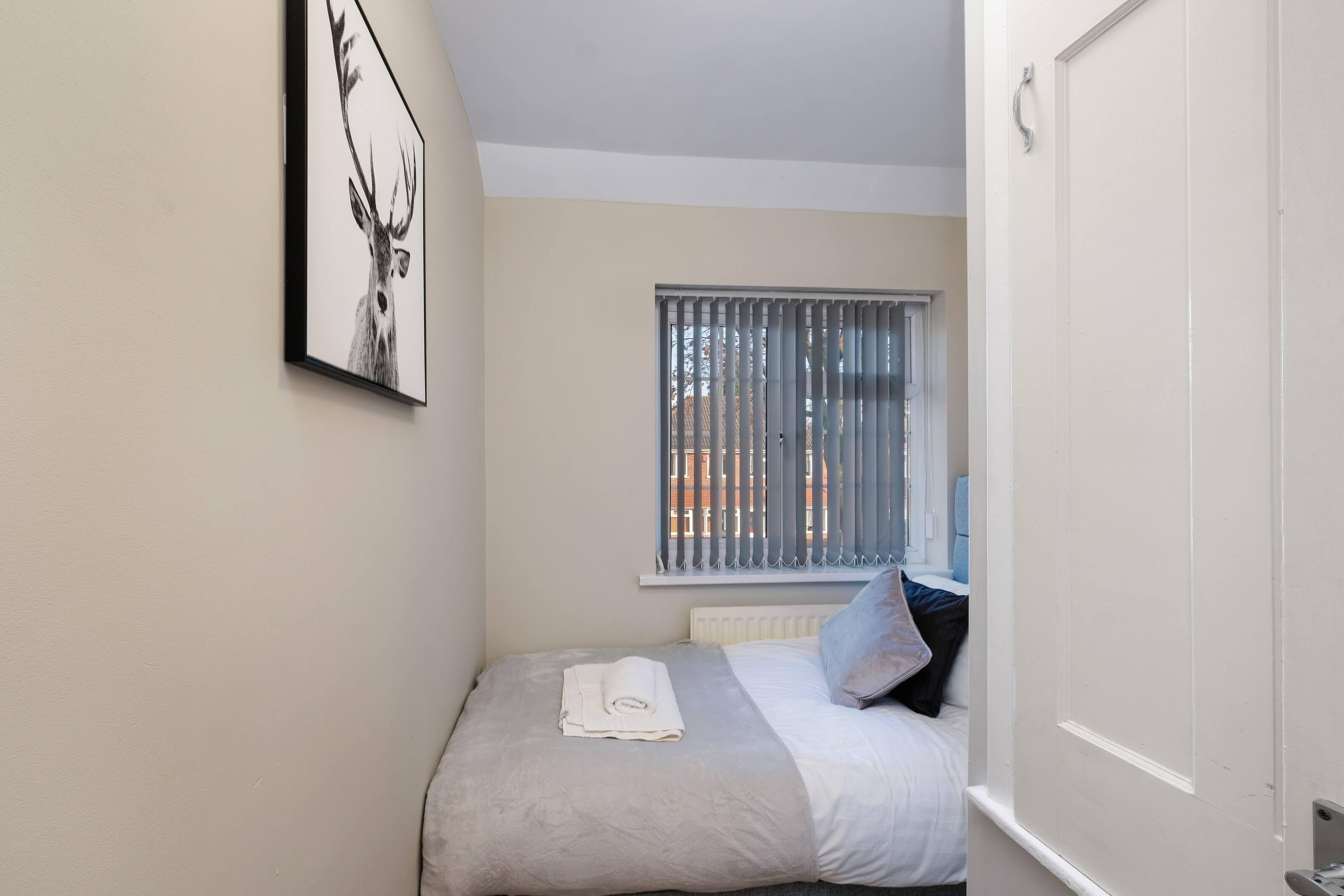 A small, neatly made bed with a folded towel, a window with blinds, and a framed picture on the wall.