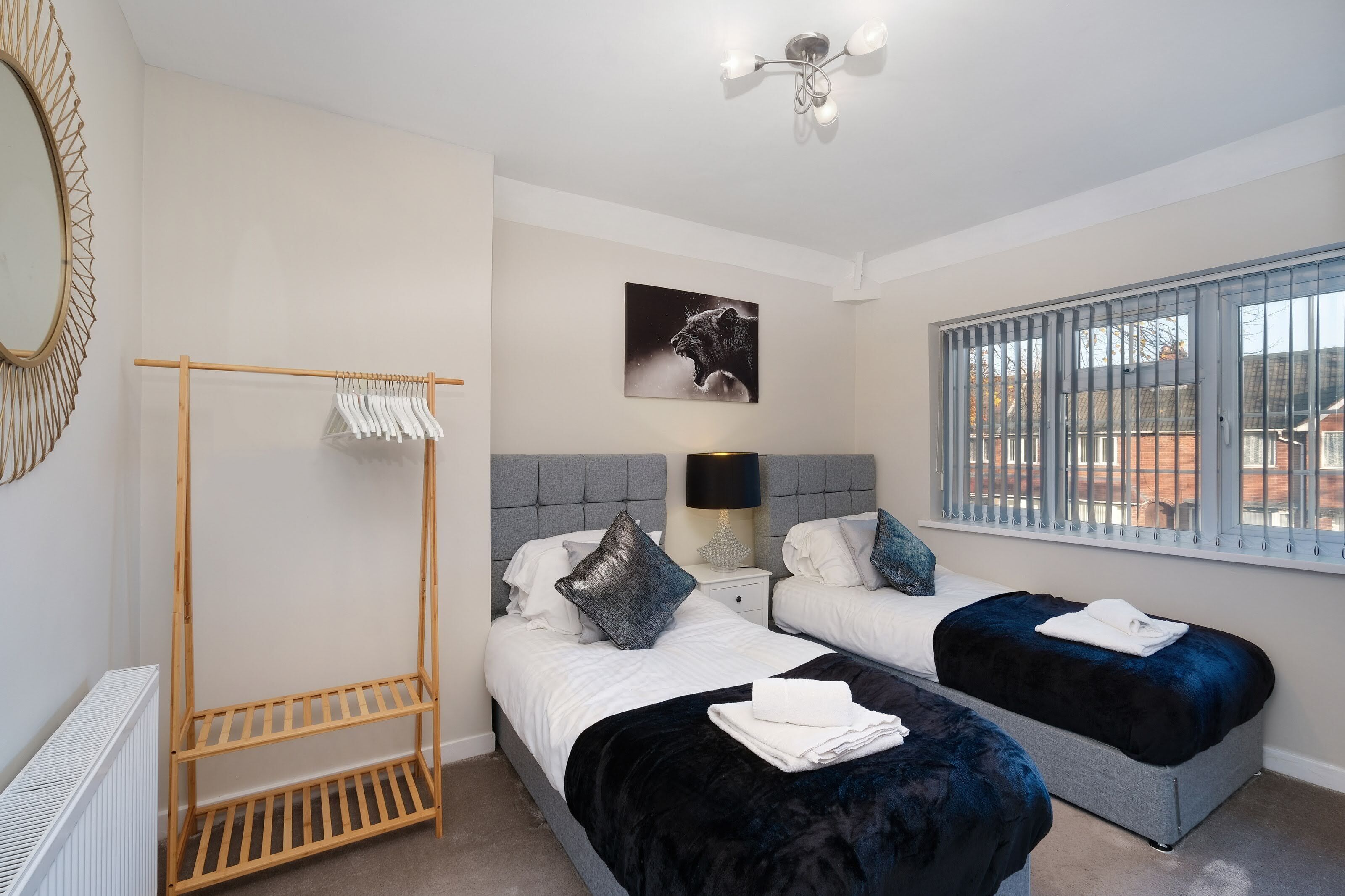 A bedroom with two beds, a wooden drying rack, a radiator, a wall-mounted mirror, a ceiling light fixture, and a window with blinds.