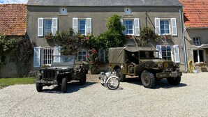Een huis met twee verdiepingen, een grindpad, geparkeerde militaire voertuigen en een fiets.