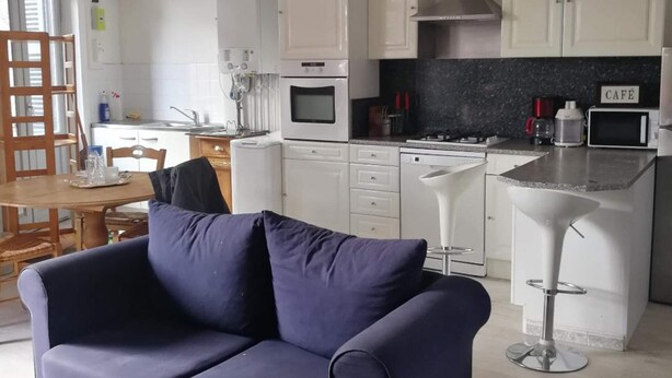 Una cocina moderna con armarios blancos, horno integrado y microondas. Hay un comedor con mesa y sillas de madera, y un sofá azul en primer plano.