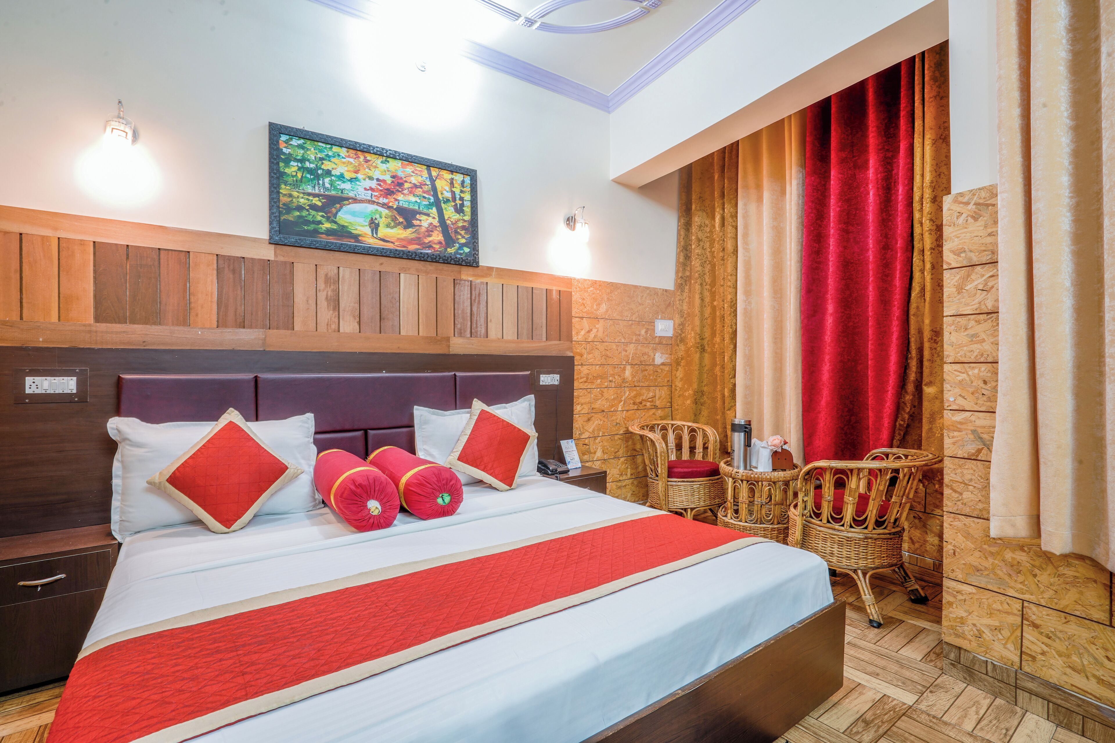 Quarto de hotel com uma cama grande, cabeceira de madeira, um quadro na parede e vista do exterior através de uma janela com cortinas vermelhas.