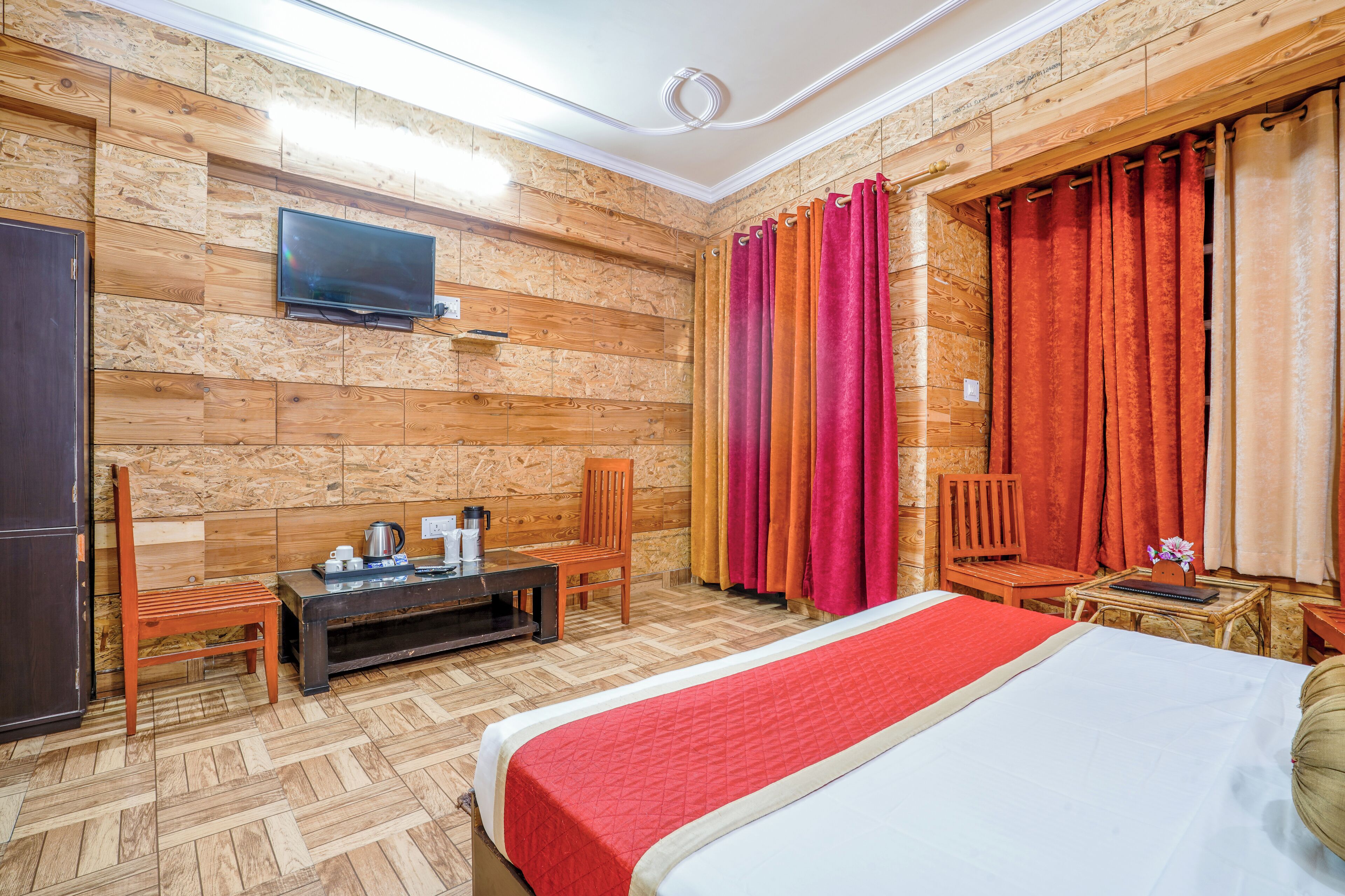 Habitación de hotel con pared de madera, una cama con manta roja, un televisor montado en la pared y una ventana con cortinas.
