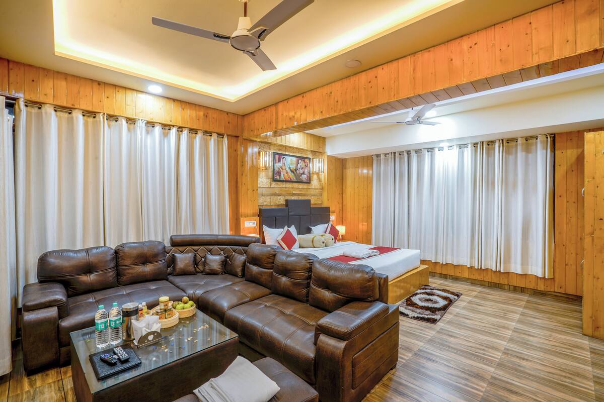 Une chambre d’hôtel moderne avec un canapé en cuir, une table basse en verre, un lit recouvert d’une courtepointe rouge et blanche, des panneaux muraux en bois et un ventilateur de plafond.