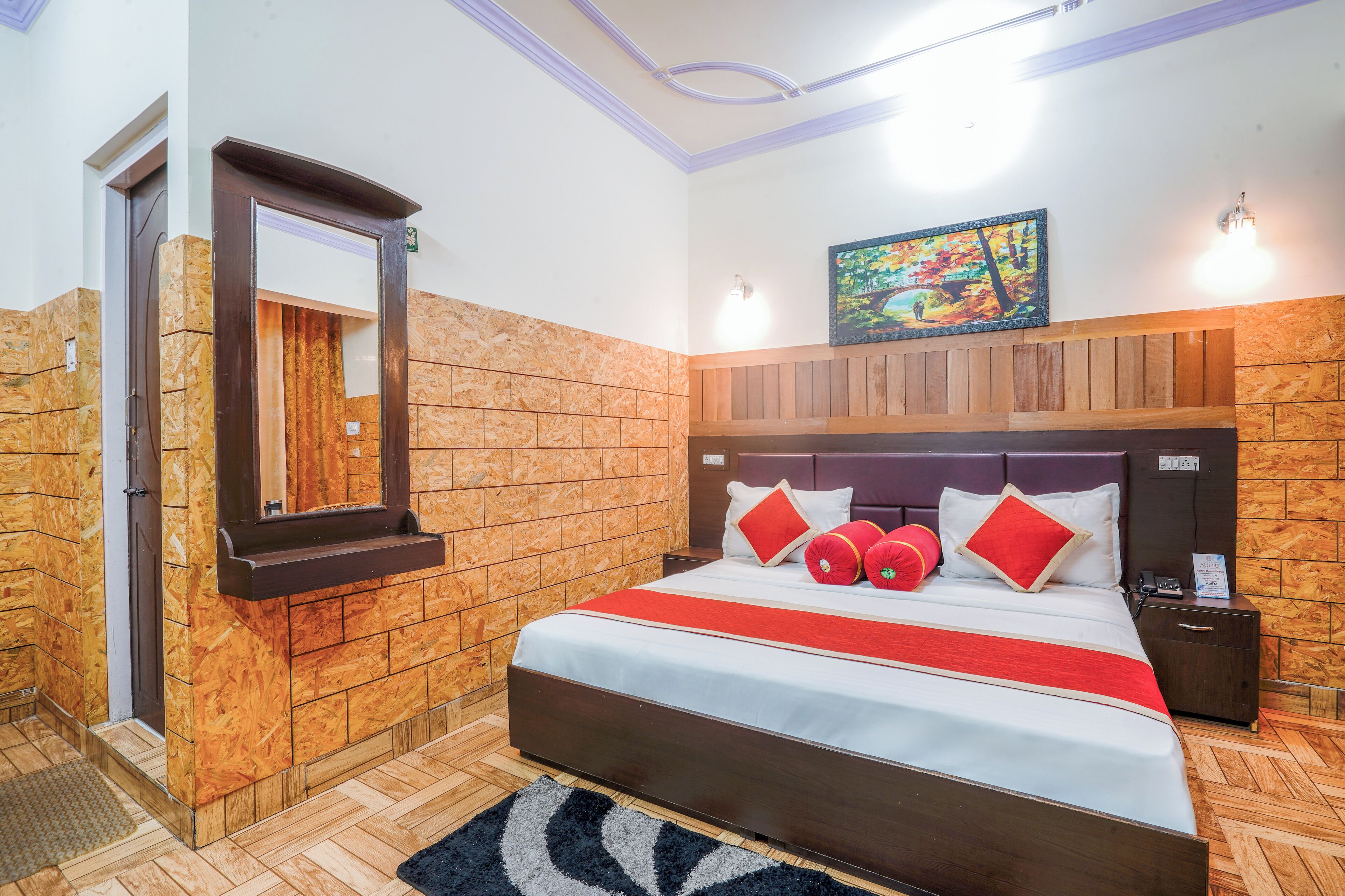 A hotel room with a wooden headboard, a large bed with red and white bedding, a wooden bench, a small table with a phone, and a painting on the wall.