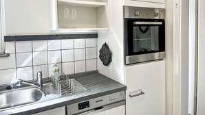 Una cuina amb un rentapeus d'acer inoxidable, un forn integrat i un armari blanc amb una cistella i gots a la repisa.