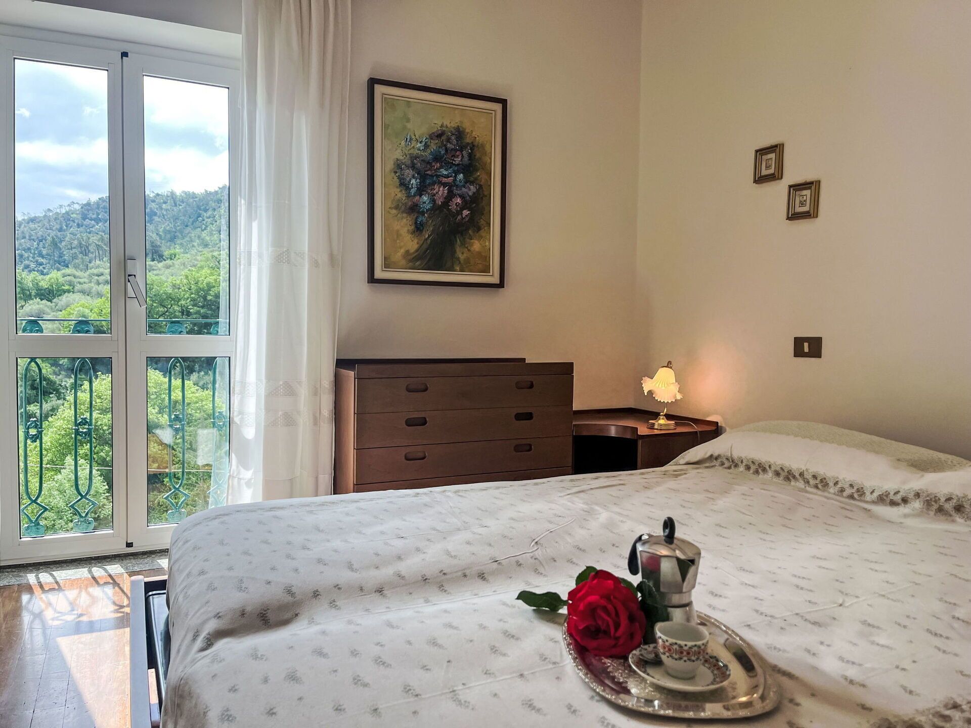 A bedroom with a bed, a dresser, a nightstand with a lamp, a framed painting, and a view of greenery through the window.