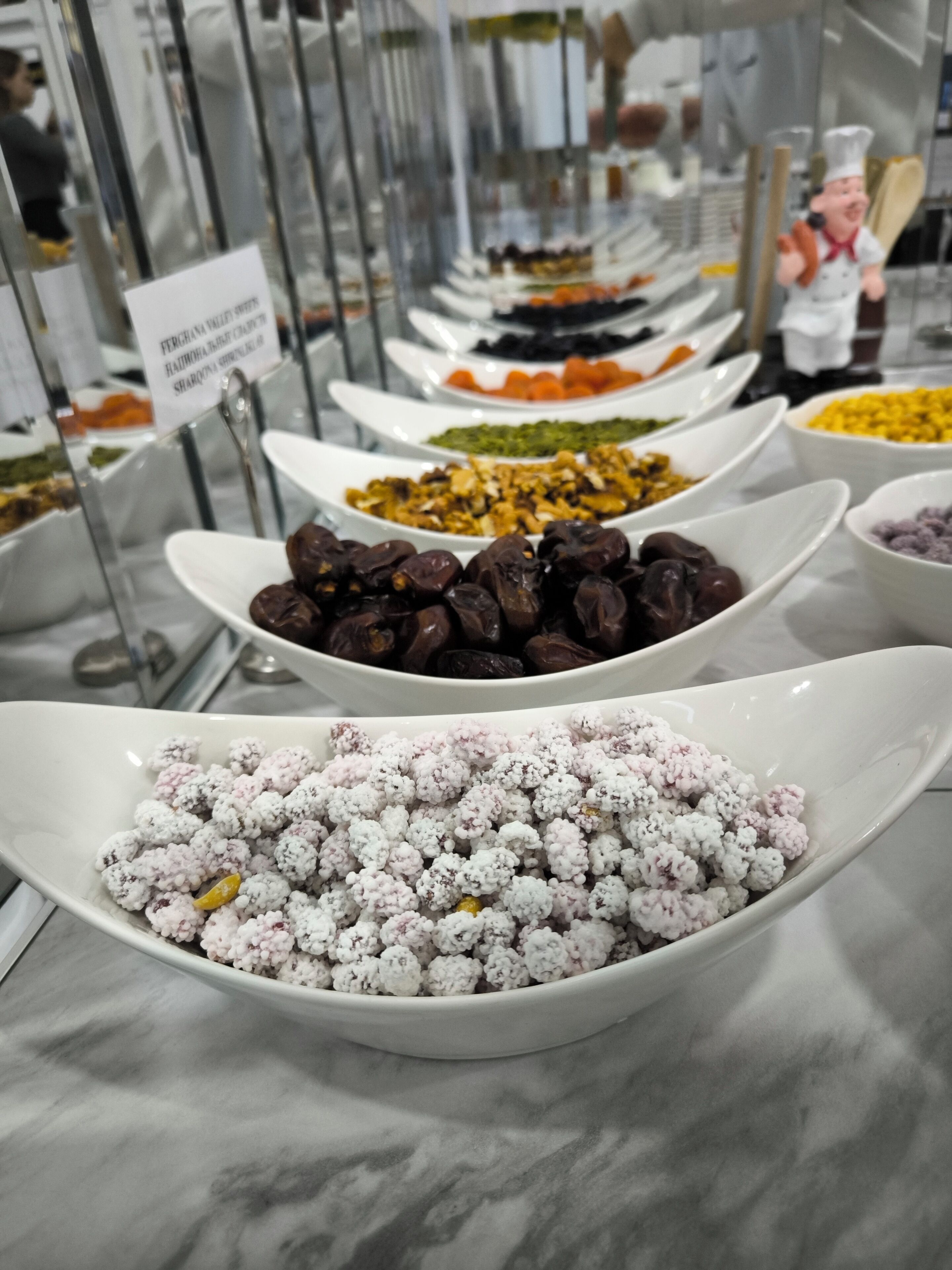 Ein Buffet mit verschiedenen Gerichten in weißen Schüsseln, darunter Datteln und ein Dessert mit rosa und weißen Toppings.