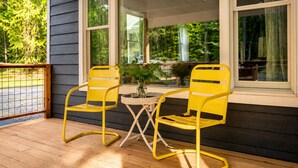 Une véranda avec deux chaises jaunes et une petite table, un plancher en bois et de grandes fenêtres donnant sur un paysage verdoyant.