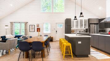 A spacious loft with a high ceiling, a dining area with a long table and chairs, a kitchen with modern appliances, and a large window providing natural light.