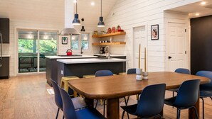 Une cuisine moderne dotée d’un îlot central, de luminaires suspendus et d’un coin repas avec une table en bois et des chaises bleues.