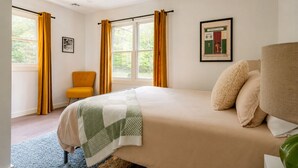 Une chambre à coucher avec un lit, une chaise, une fenêtre avec des rideaux et un tableau encadré au mur.