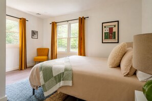 A bedroom with a bed, a chair, a window with curtains, and a framed picture on the wall.