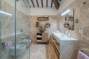 A modern bathroom with a glass shower enclosure, a bidet, a vanity with a mirror, and wooden shelves with toiletries.