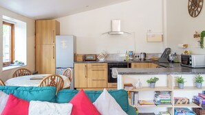 Une cuisine compacte avec des meubles en bois, un four encastré et un four à micro-ondes. On y trouve un coin repas avec une table et des chaises, ainsi qu’une bibliothèque remplie de livres.