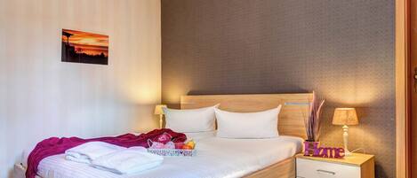 Un lit bien fait, avec du linge de lit blanc et une couverture rose, une table de chevet avec une lampe, et un petit objet décoratif sur lequel est inscrit le mot « Accueil ».