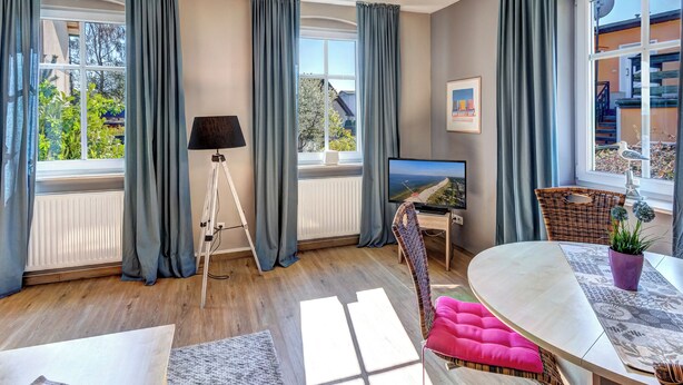 Ett modernt vardagsrum med stora fönster, en platt-TV, en golvlampa och en matplats med ett bord och stolar.