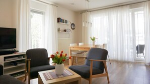 A living room with a flat-screen TV, two armchairs, a coffee table with a vase of flowers, a bookshelf, and a dining area with a table and chairs.