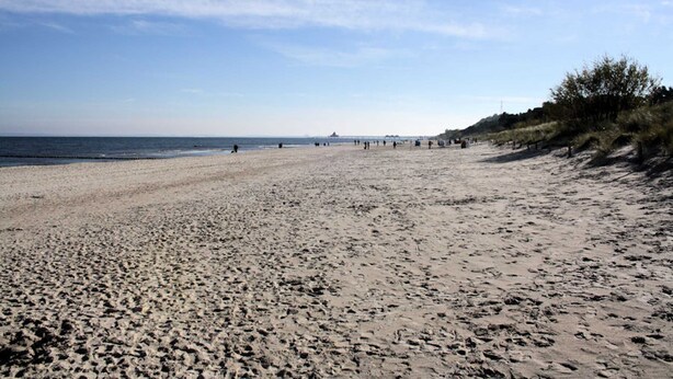 A wide sandy beach with footprints, a clear sky, and distant structures.