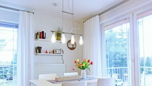 A bright, modern dining area with a white table, two chairs, a vase with flowers, a bookshelf with books and a radio, and a view of the outdoors through large windows.