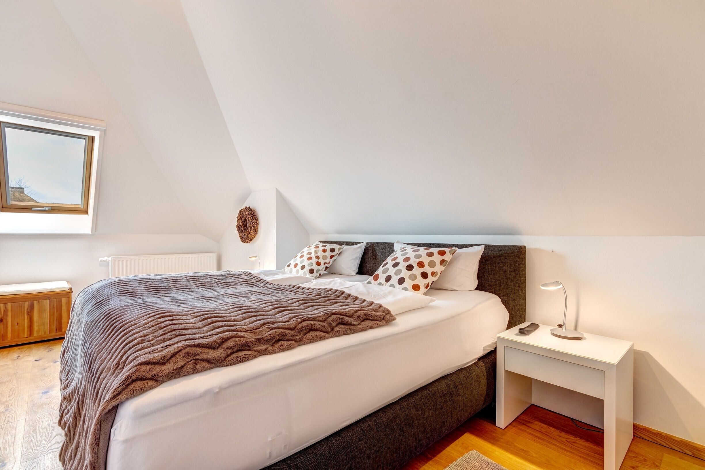 A bedroom with a large bed, a nightstand with a lamp, a radiator, and a window with a view of the sky.