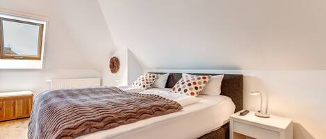 A bedroom with a large bed, a nightstand with a lamp, a radiator, and a window with a view of the sky.
