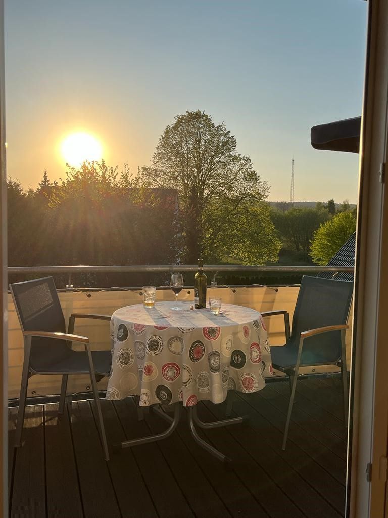 Ein runder Tisch mit gemustertem Tischläufer, vier Stühle und einer Weinflasche auf einem Balkon mit Blick auf Sonnenuntergang und Bäume.