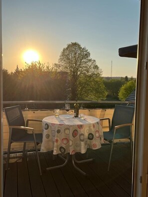 Een ronde tafel met een tafelkleed met patroon, vier stoelen en een fles wijn op een balkon met uitzicht op de zonsondergang en bomen.