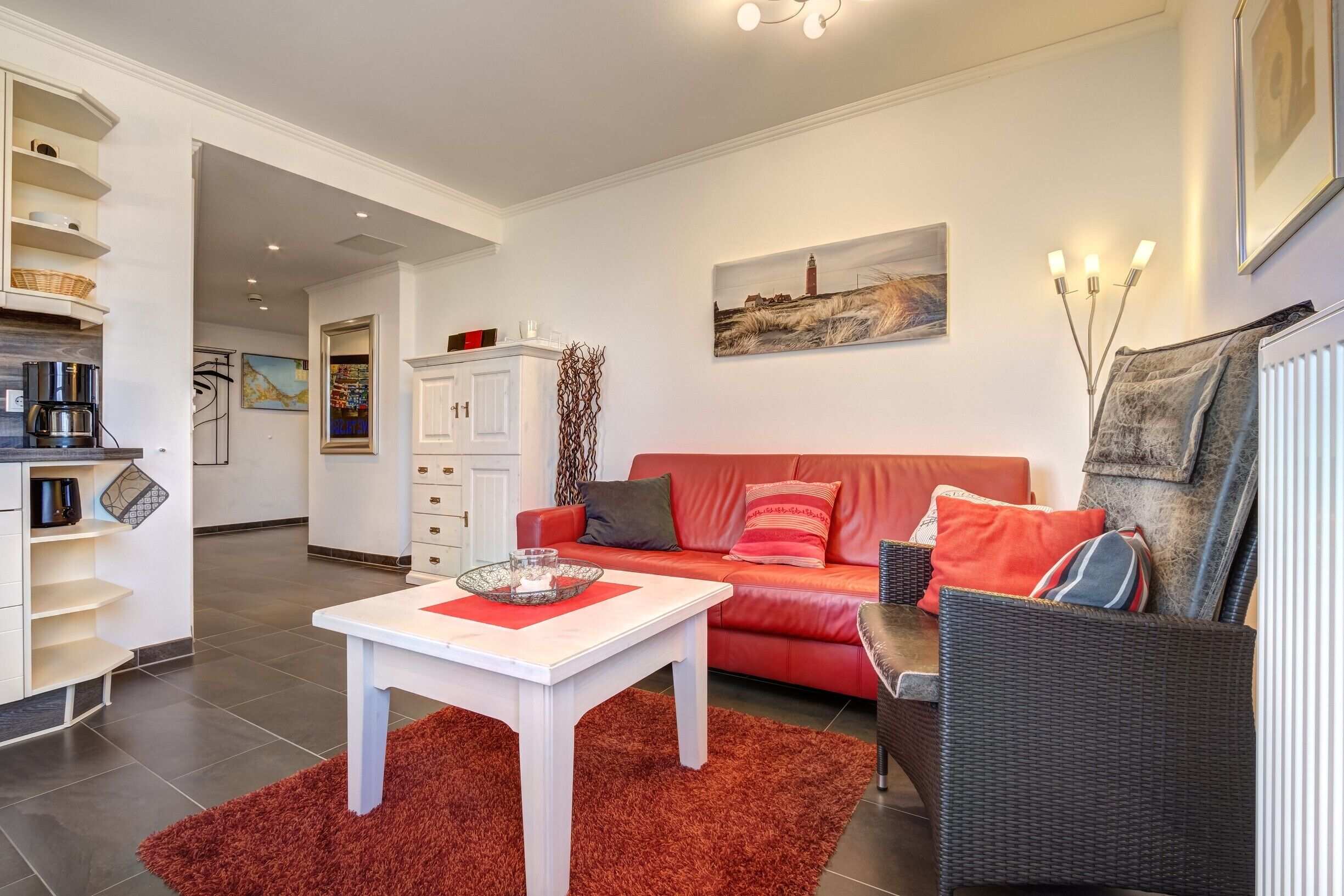 Uma sala de estar moderna com um sofá vermelho, mesa de centro branca e uma cadeira de vime. Há uma área de cozinha com armários brancos e uma máquina de café.