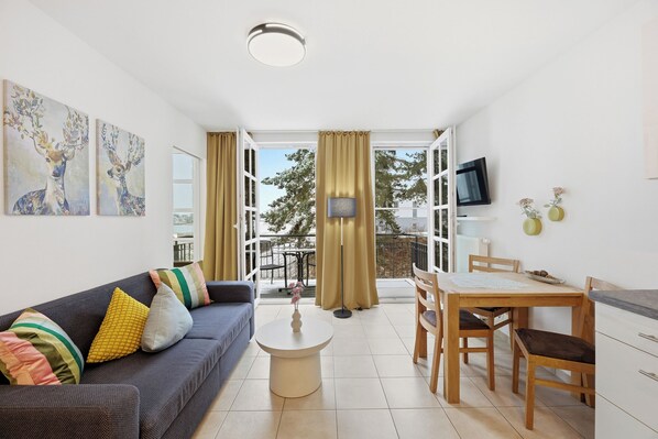 A modern living room with a sofa, coffee table, and dining area. There are two paintings on the wall, a TV mounted above the dining table, and a balcony with curtains.