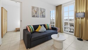 Un salón moderno con un sofá gris oscuro, cojines coloridos, una mesa de centro blanca y un amplio ventanal con cortinas.