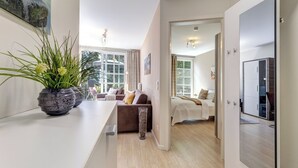 Ein modernes Wohnzimmer mit einer Couch, einem Couchtisch und einer Topfpflanze. Ein Bett mit weißer Bettdecke und ein an der Wand montierter Fernseher sind ebenfalls vorhanden.