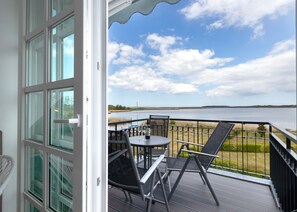 A balcony with a table and chairs, overlooking a body of water and a distant shoreline. - Residenz am Balmer See Whg. 65 with lake view, sauna & wellness by the water (Balm)