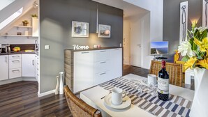 Uma cozinha moderna com armários brancos, uma área de refeições com mesa posta para dois e uma sala de estar com sofá e televisão.