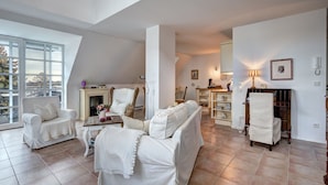 A spacious living room with a high ceiling, white furniture, a fireplace, and a view through large windows.
