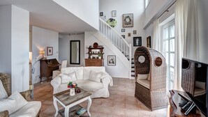 A spacious living room with a white sofa, a wooden coffee table, and a wicker chair. A staircase with a wooden railing leads to an upper level.