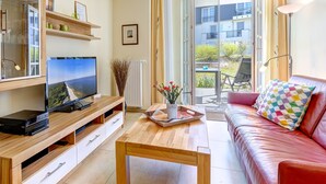 A modern living room with a wooden TV stand, a flat-screen TV, a wooden coffee table, a red leather sofa, and a balcony with a view of buildings and greenery.